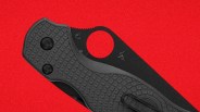 Black folding knife with textured handle and circular thumb hole on a red background.