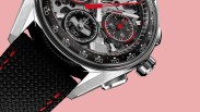 Close-up of a silver chronograph watch with black and red details and a black fabric strap with red stitching on a pink background.