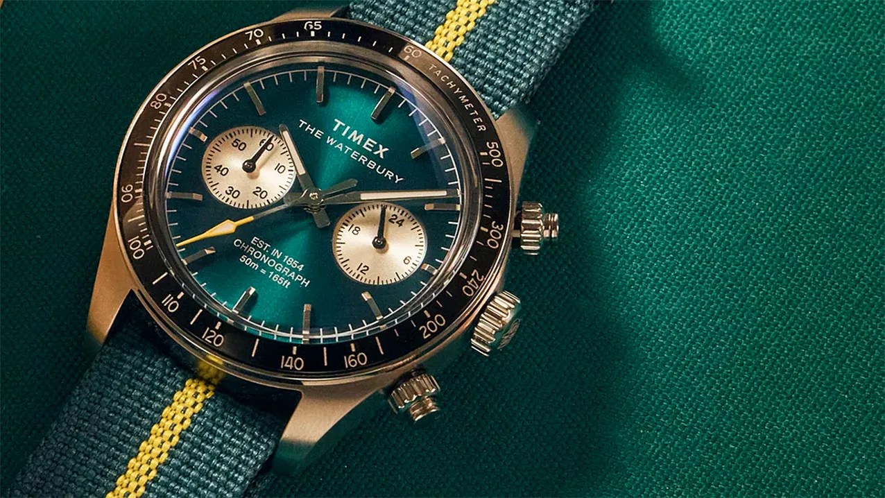 Timex Waterbury chronograph watch with teal dial, black tachymeter bezel, and blue fabric strap with yellow stripe.