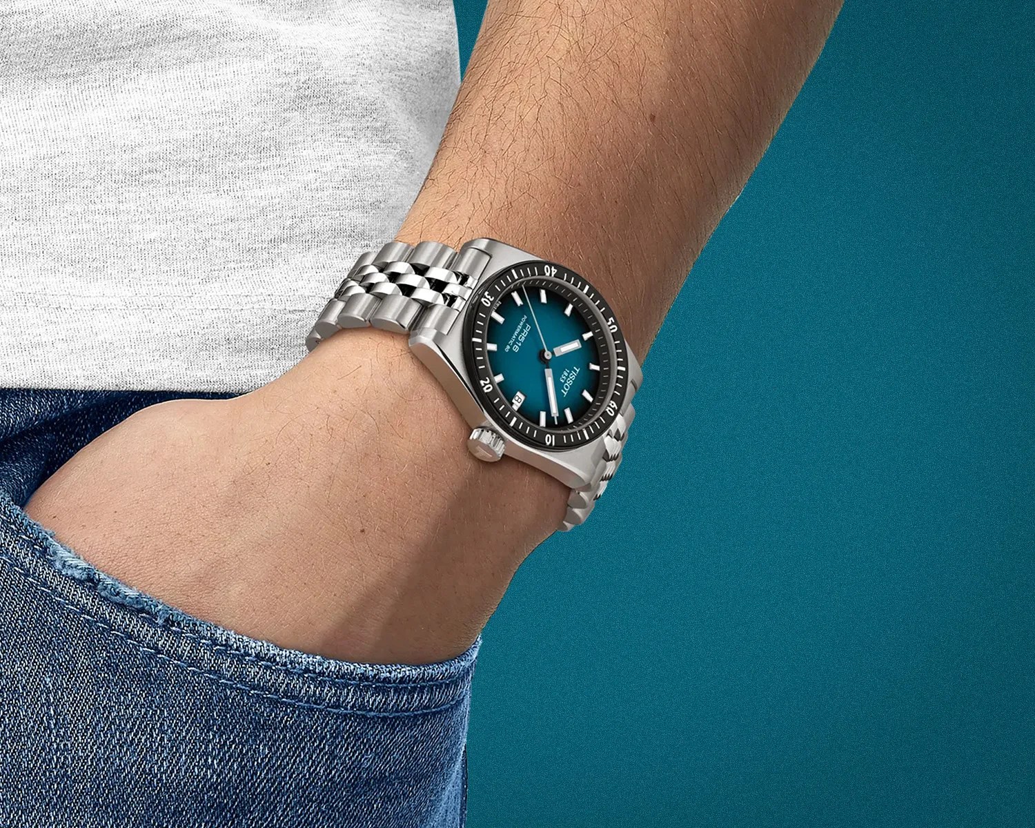 Silver metal wristwatch with a blue dial and black bezel worn on a wrist with a hand in a blue denim pocket.