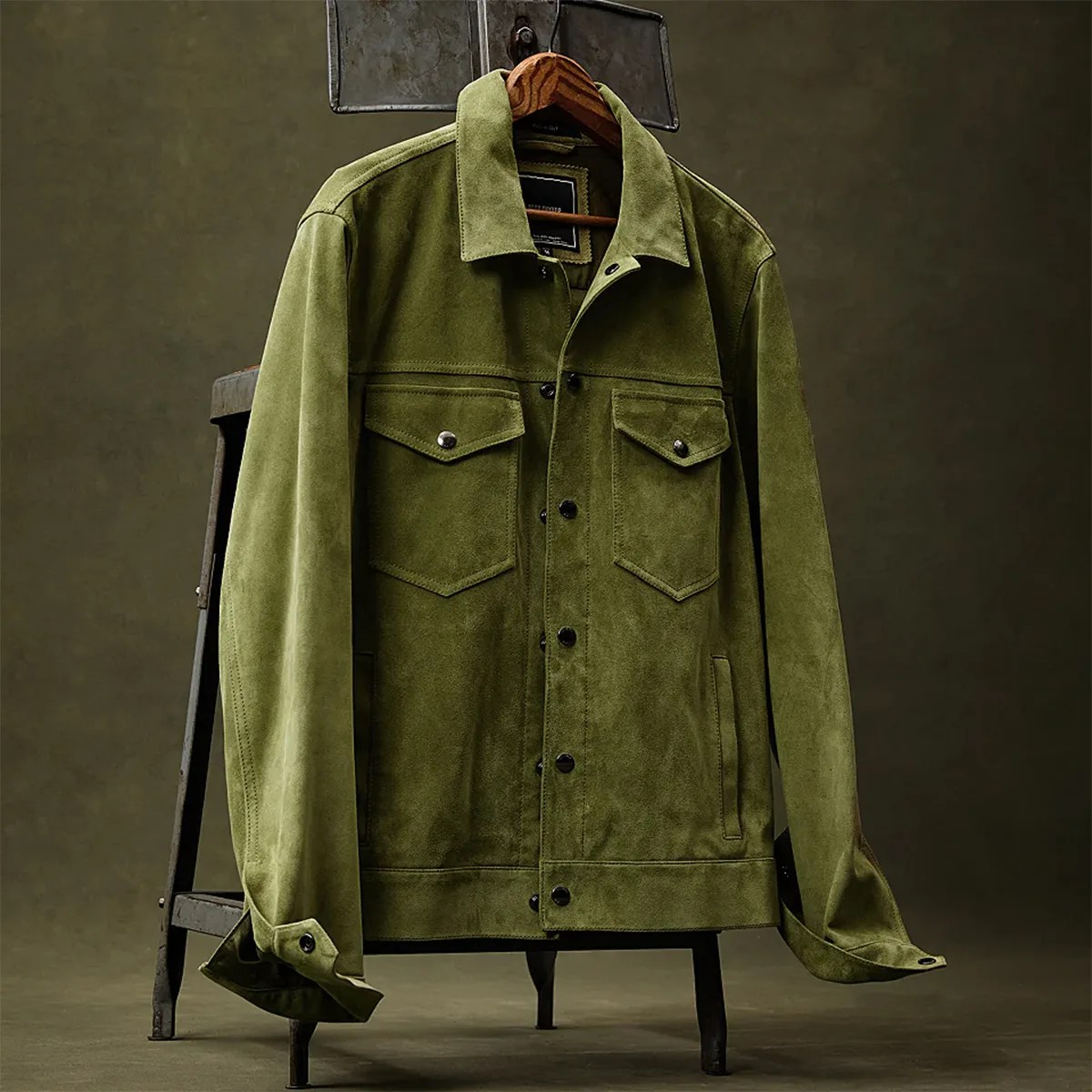 Olive green suede jacket with buttoned flap chest pockets hanging on a wooden hanger.