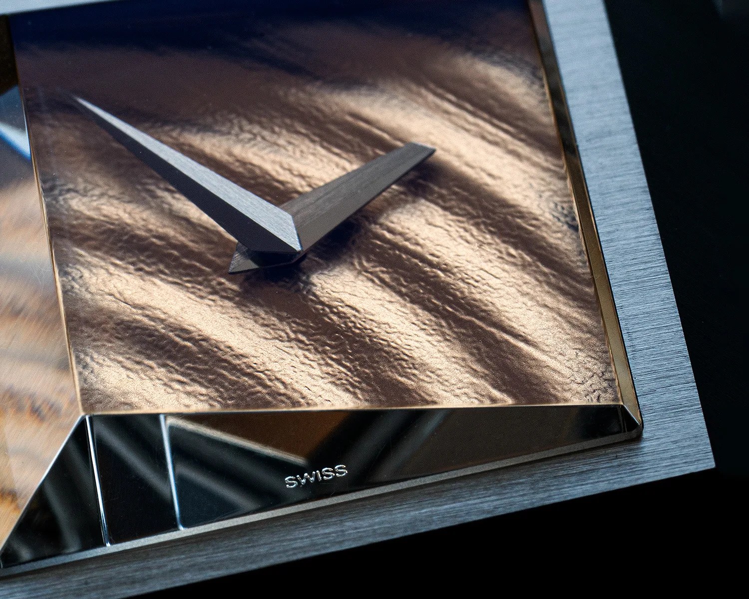 Close-up of a modern clock face with textured bronze surface, silver hands, and "SWISS" engraved on a reflective metal base.