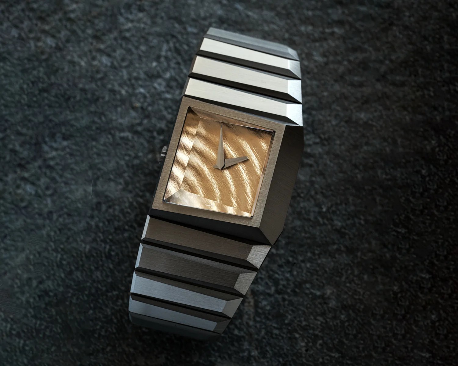 Brushed metal wristwatch with a textured gold square dial and no numerals on a dark textured surface.