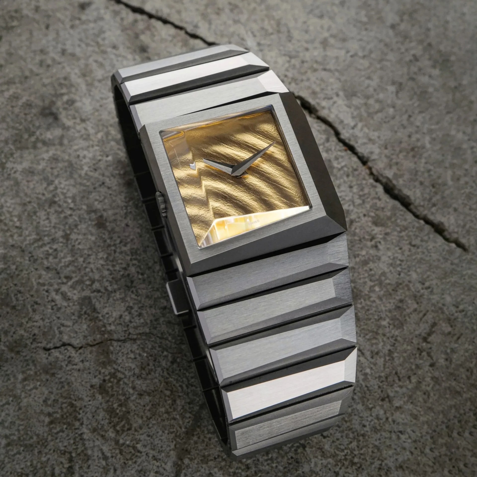 Rectangular silver metal wristwatch with a textured gold dial and no numerals on a cracked concrete surface.