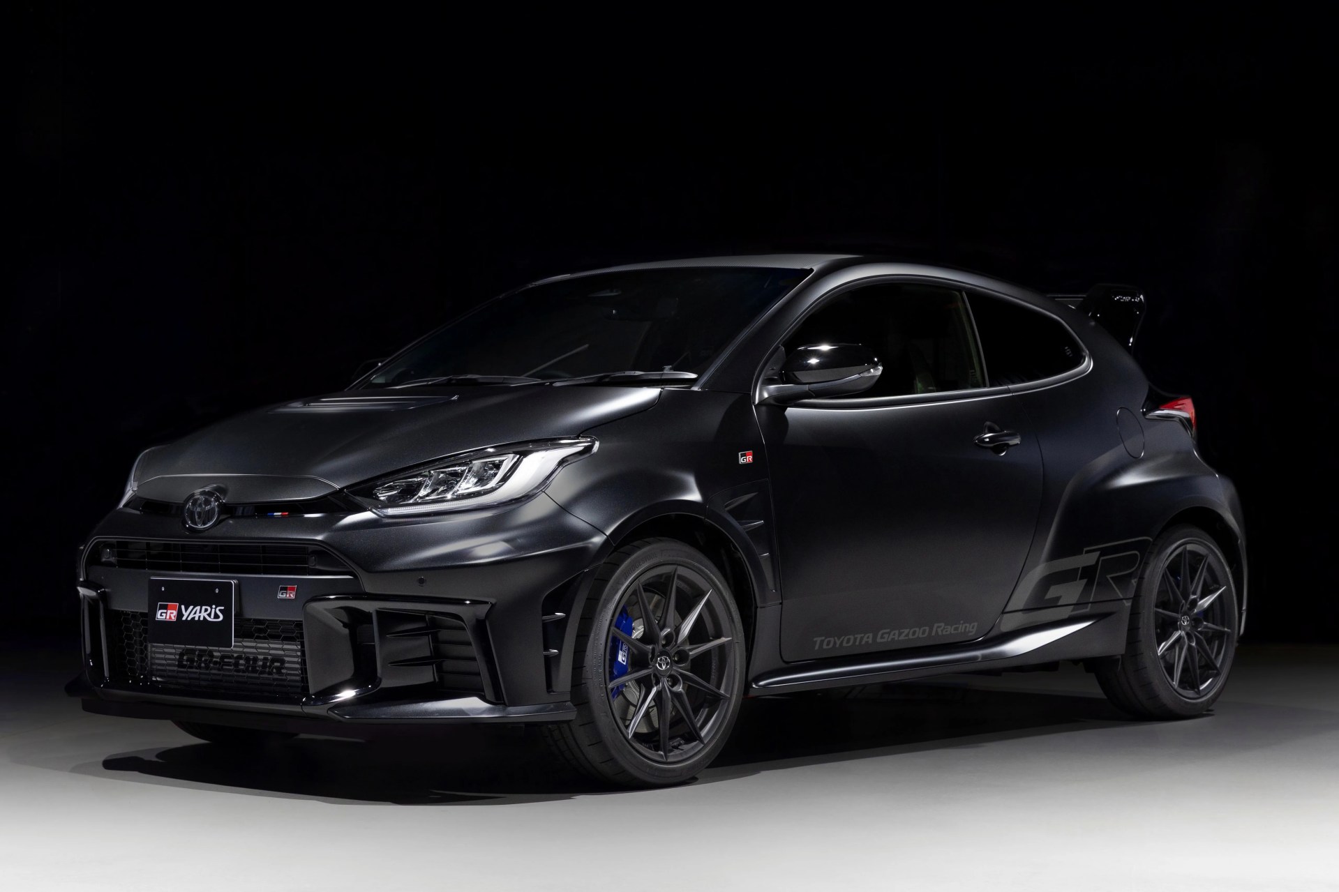 Matte black Toyota GR Yaris hatchback with sporty body kit and blue brake calipers on a gray floor.