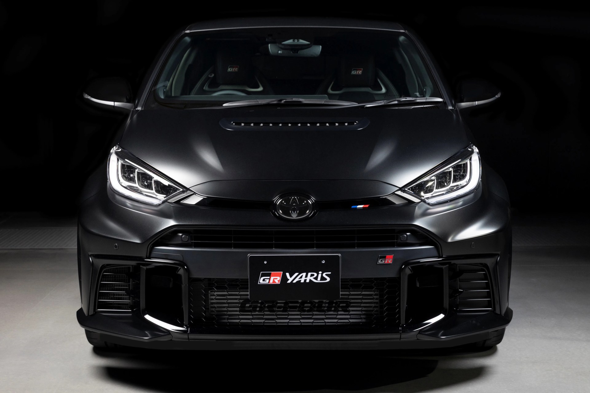 Front view of a matte black Toyota GR Yaris with LED headlights and sporty grille in a dark setting.