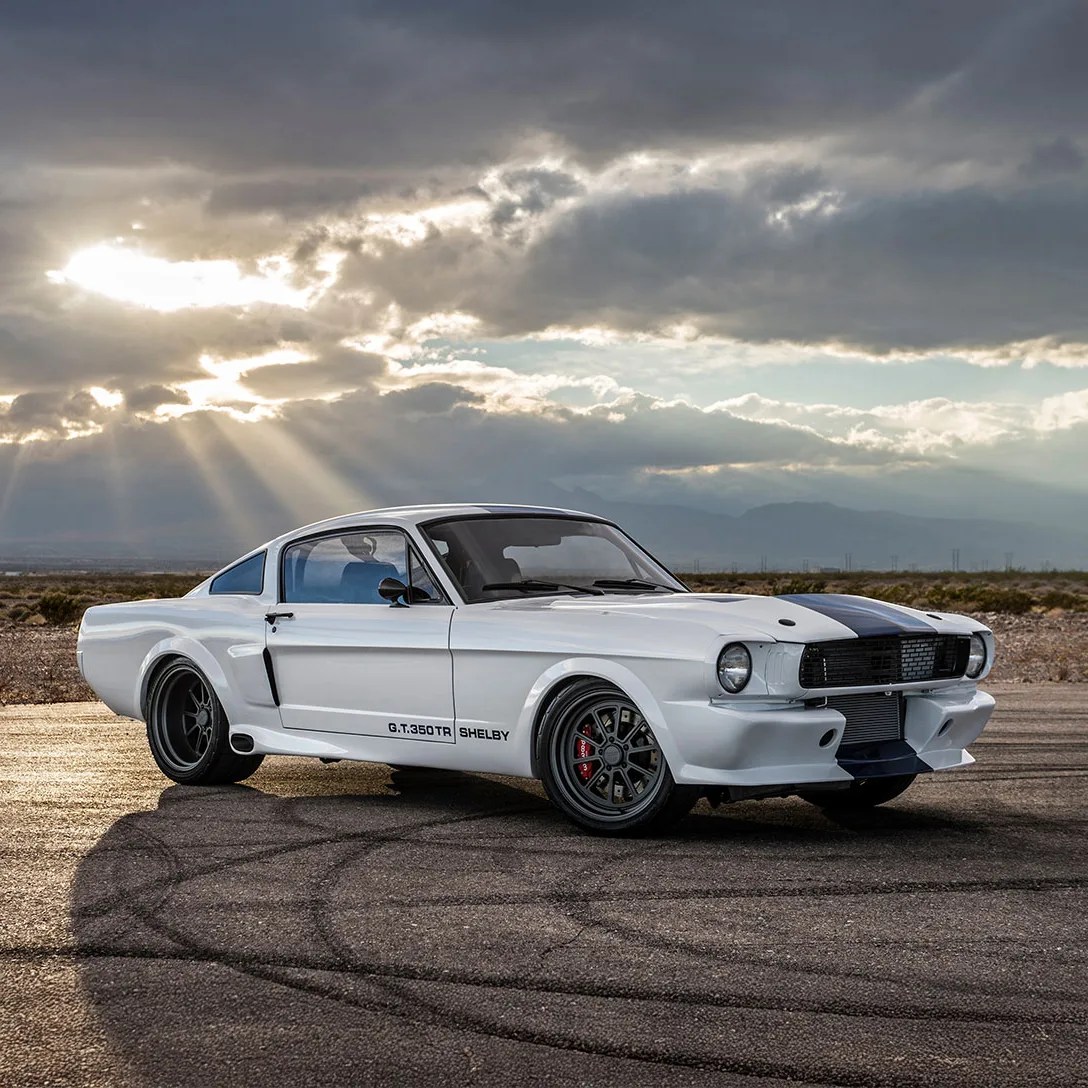 White Shelby G.T.350TR classic car with black racing stripes parked on asphalt under a cloudy sky with sun rays.