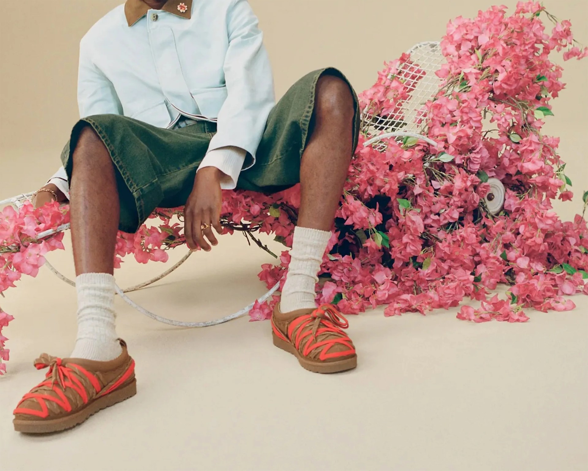 Person wearing olive green shorts, white socks, and brown sandals with bright orange straps sitting on a white metal chair covered in pink flowers.