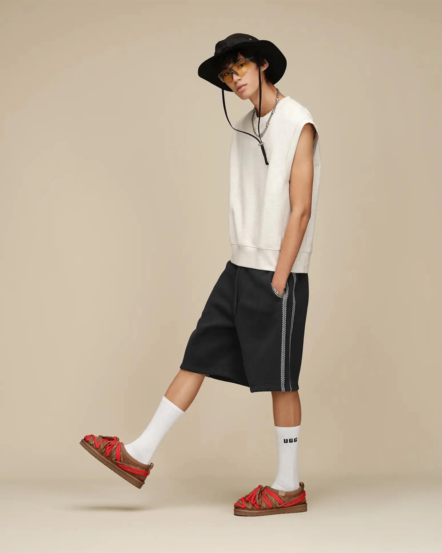 Person wearing a sleeveless beige top, black shorts with side stripes, white socks, brown sandals with red straps, black wide-brim hat, and yellow-tinted glasses.