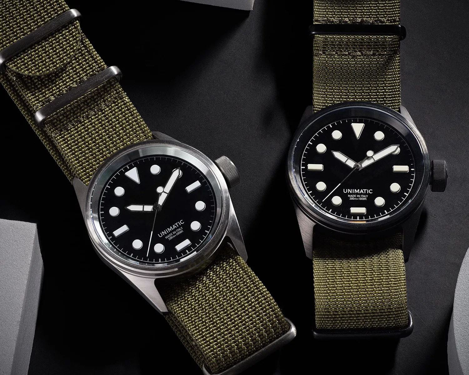Two Unimatic wristwatches with black dials and olive green fabric straps on a dark surface.