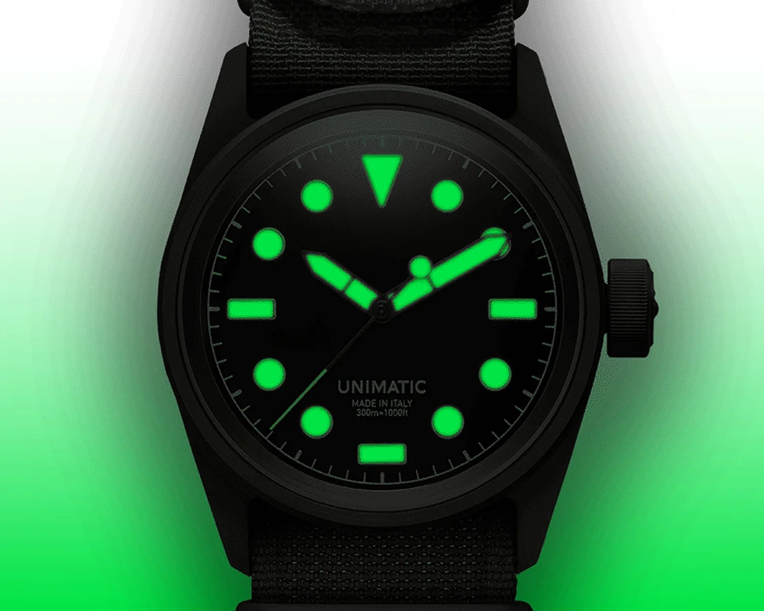 Black Unimatic watch with glowing green hour markers and hands on a black fabric strap.