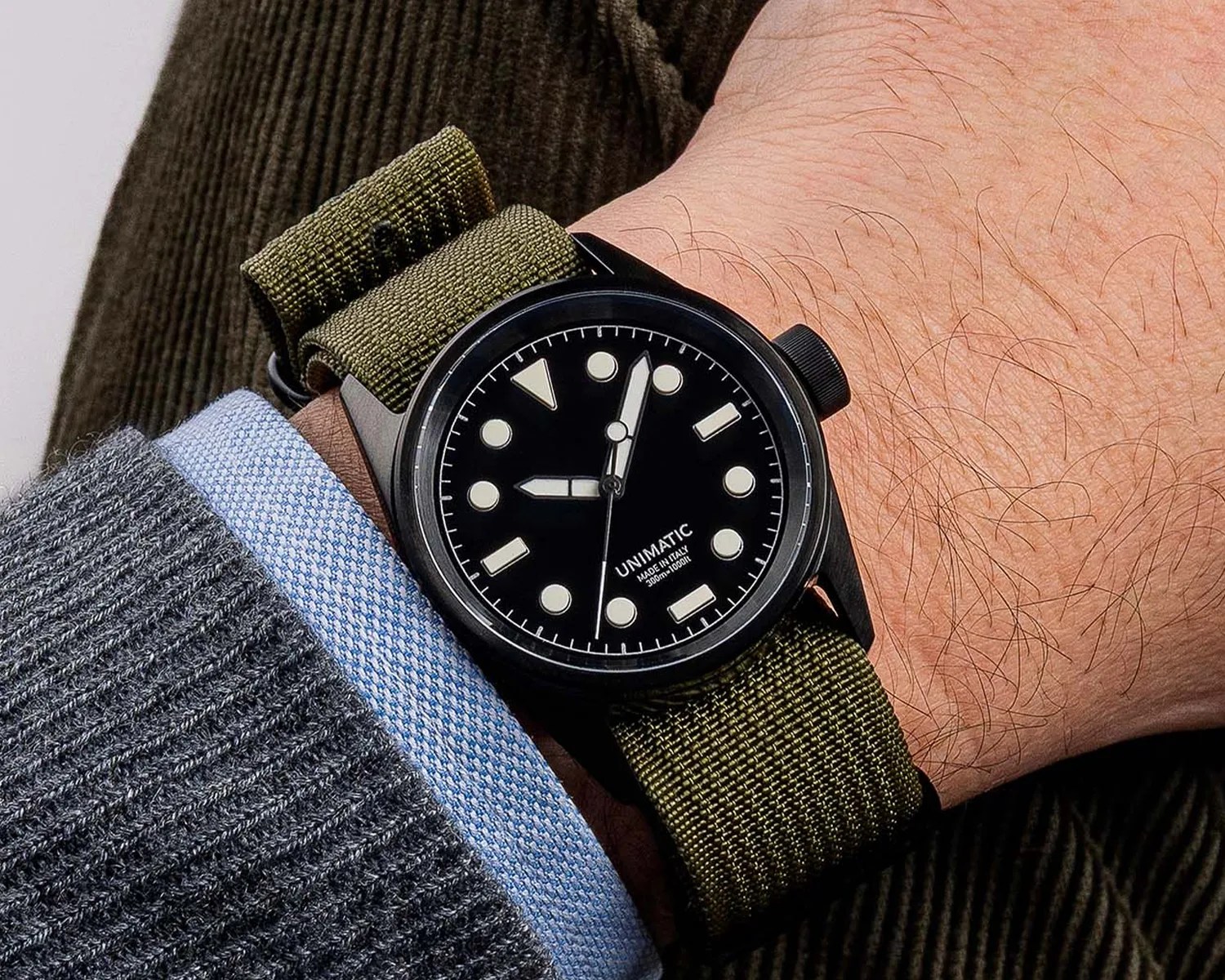 Black Unimatic wristwatch with luminous markers and hands on an olive green fabric strap worn over a blue shirt and gray sweater.