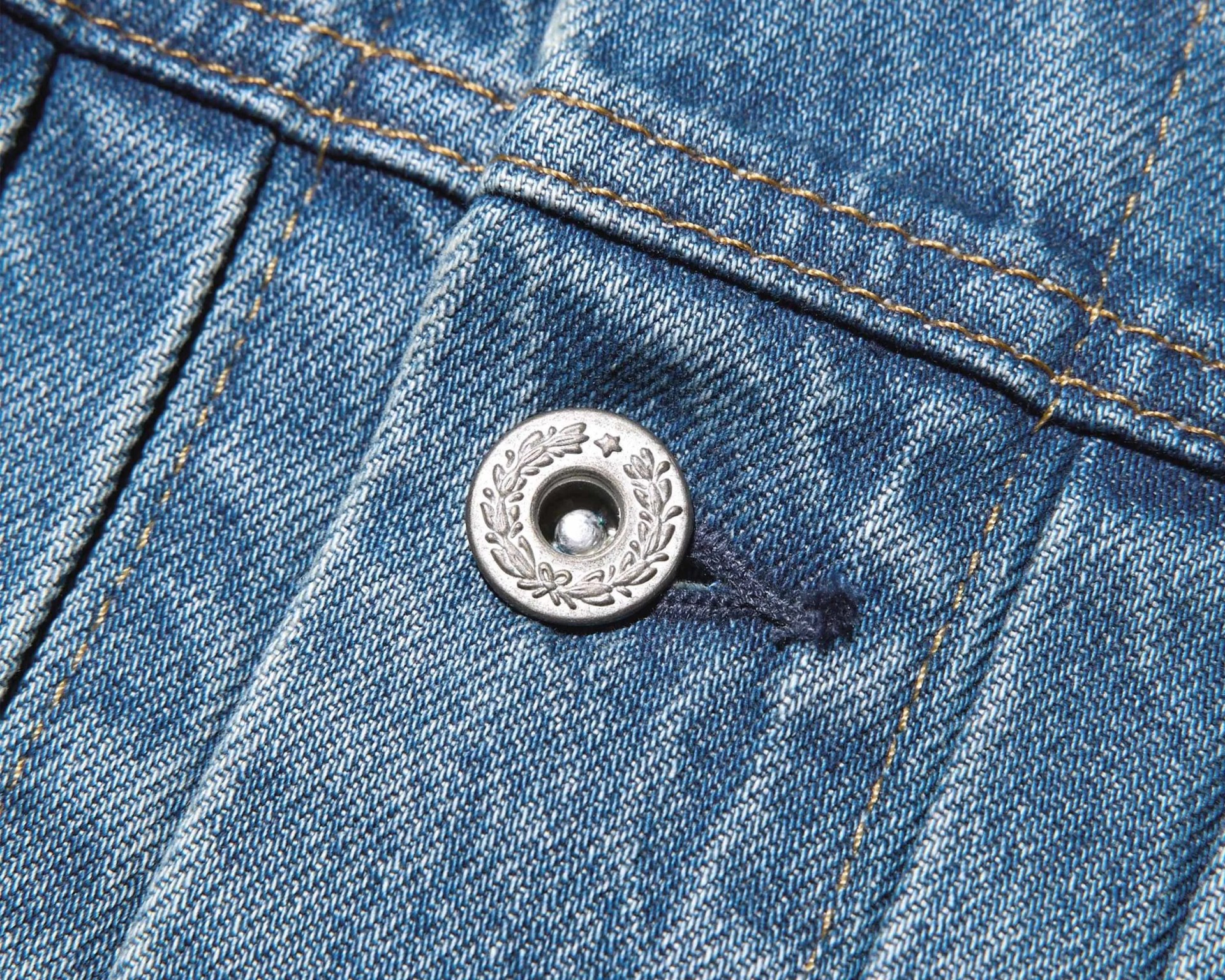 Close-up of a silver decorative button with a leaf and star design on blue denim fabric with orange stitching.