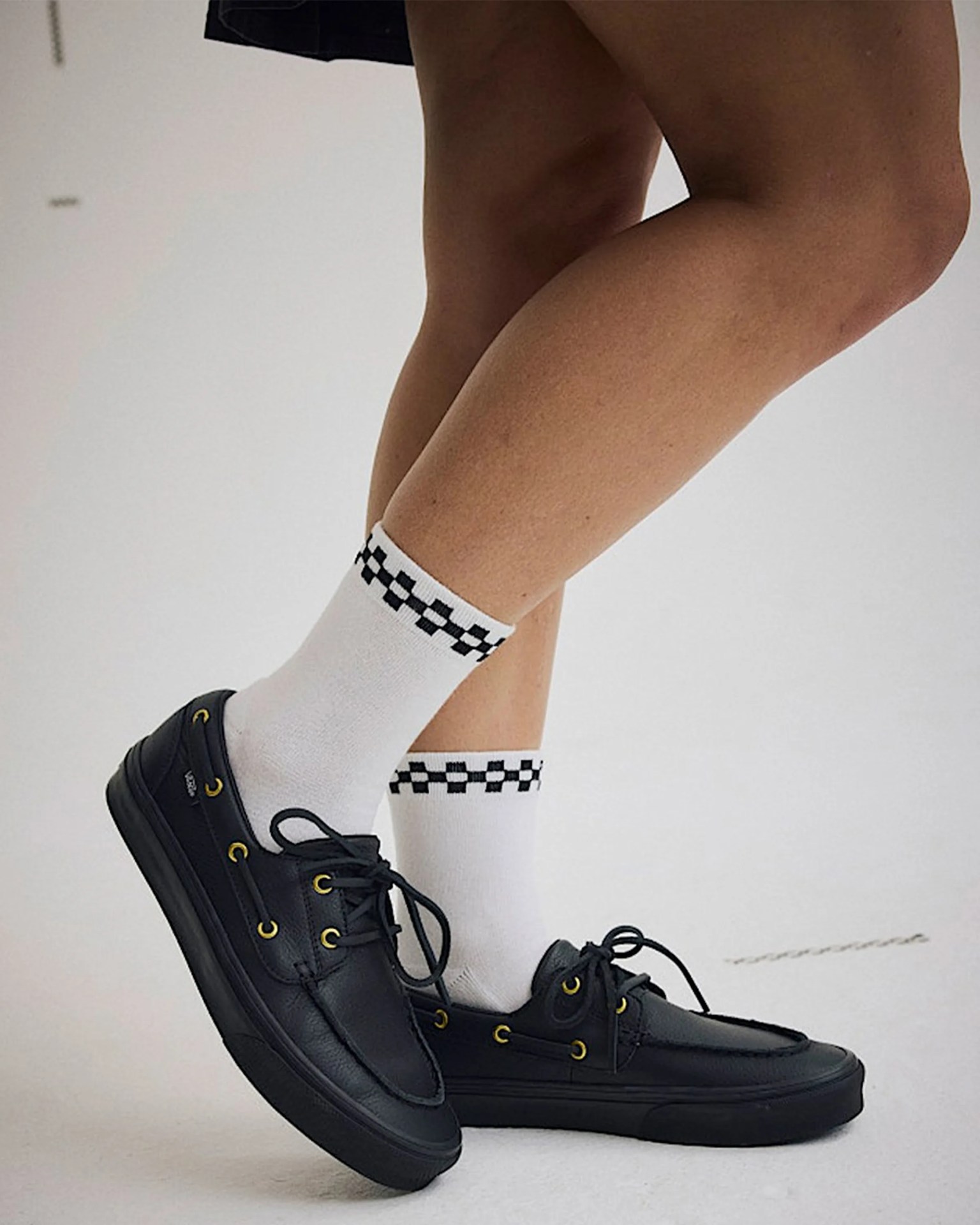 Black leather boat shoes with yellow eyelets worn with white socks featuring a black checkerboard pattern near the top.