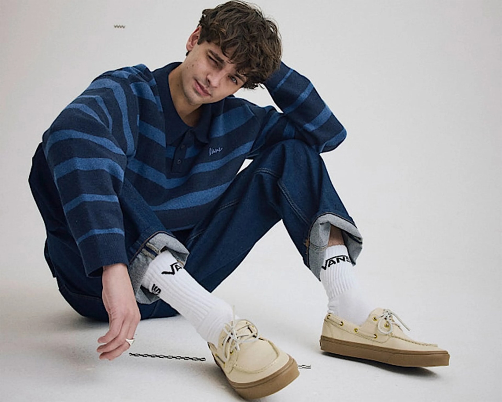 Person sitting on the floor wearing a blue striped sweater, cuffed dark blue jeans, white Vans socks, and beige boat shoes.