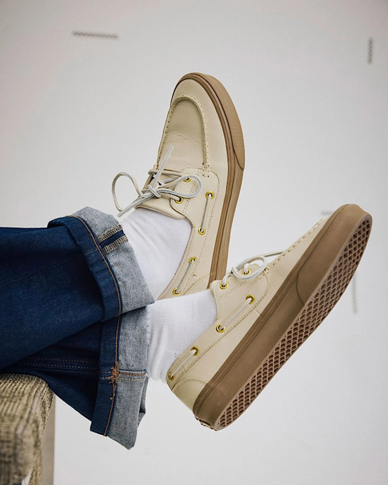 Beige leather boat shoes with white laces and brown rubber soles worn with white socks and cuffed blue jeans.