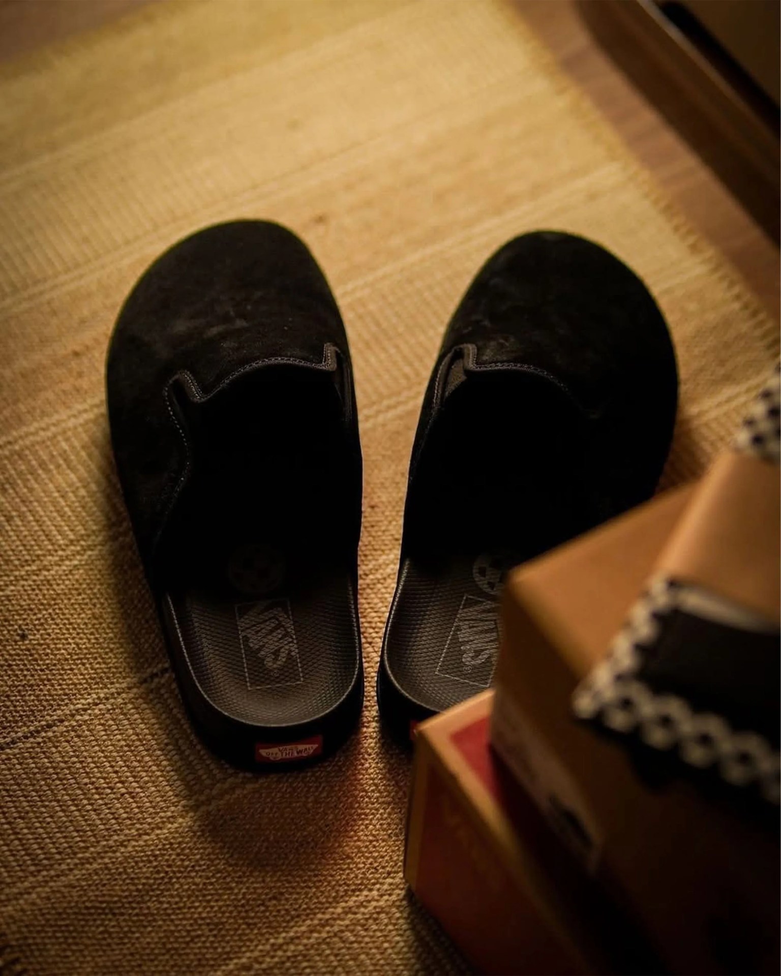 Pair of black Vans slip-on shoes on a textured beige rug next to stacked shoe boxes.