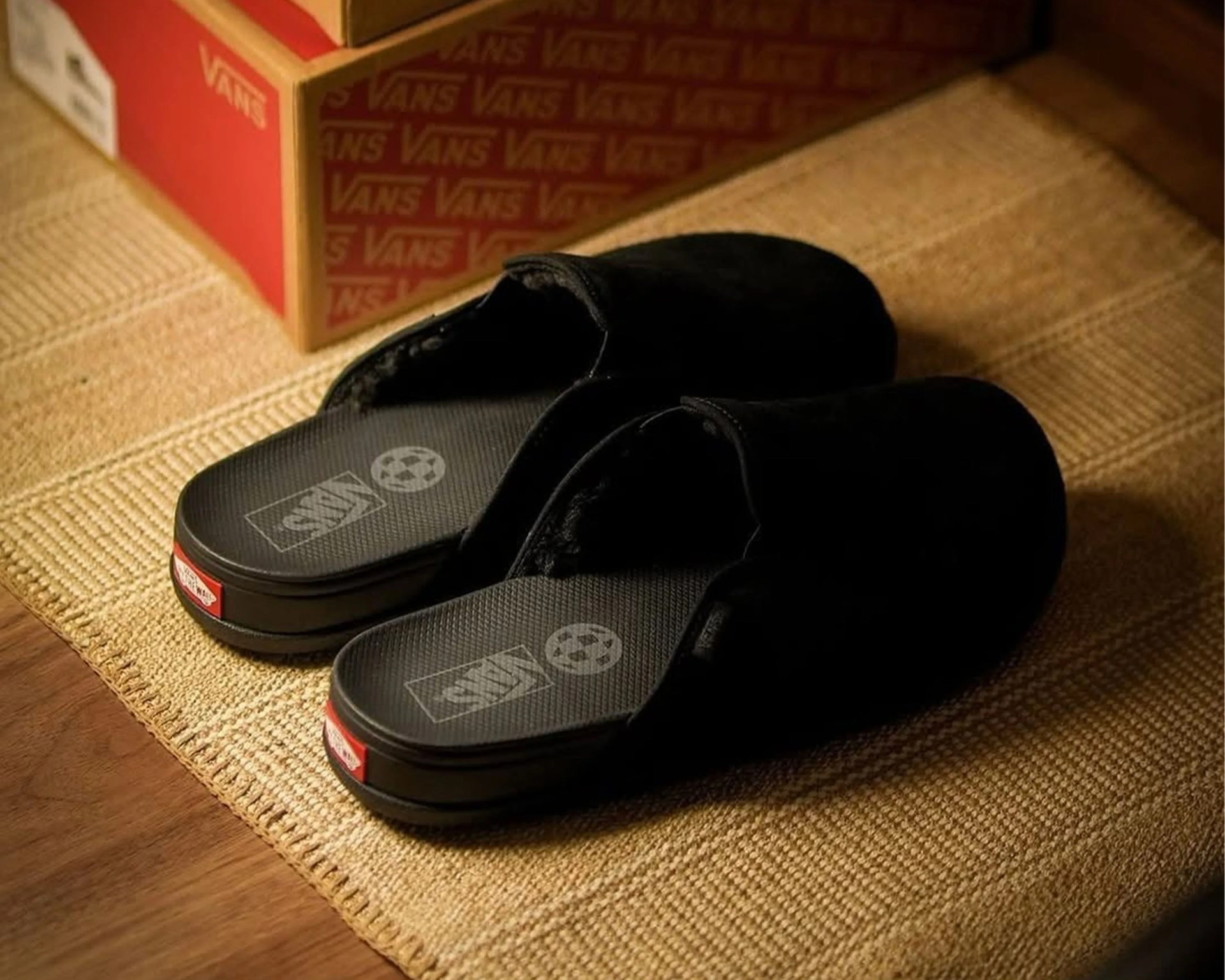Pair of black Vans slide slippers on a woven mat with Vans shoe boxes in the background.