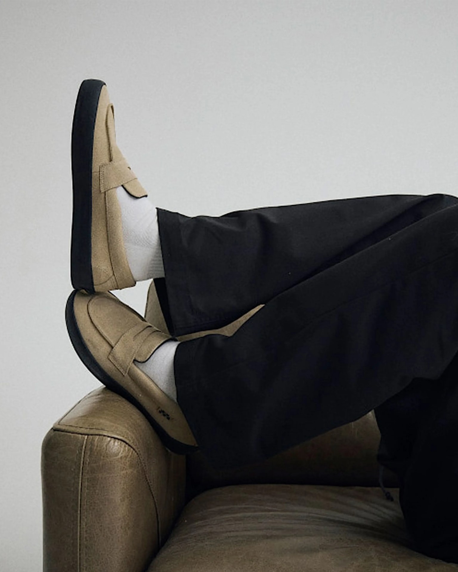 Beige suede loafers with black soles worn with white socks and black pants resting on a brown leather couch.