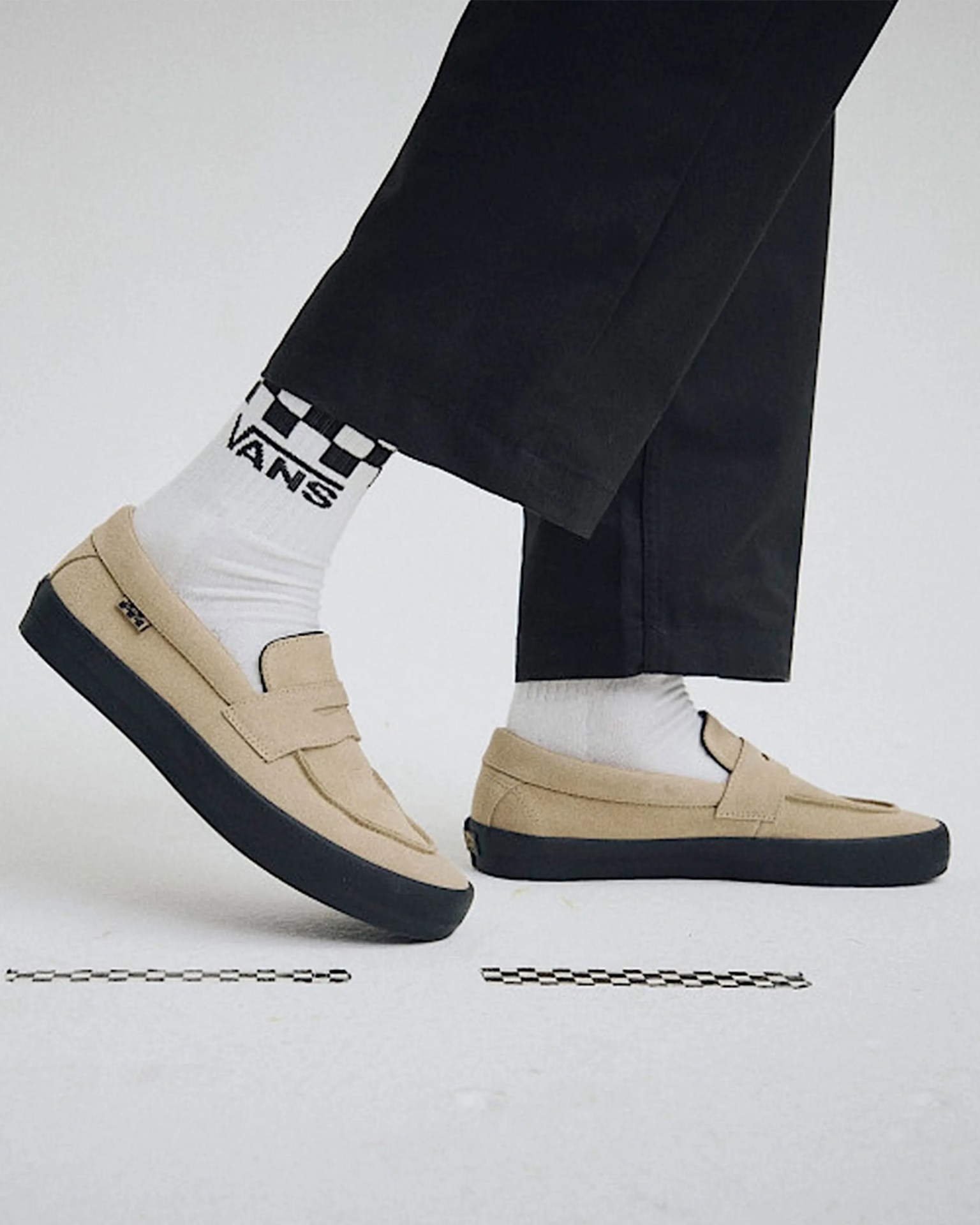 Beige suede loafers with black rubber soles worn with white Vans socks and black pants.