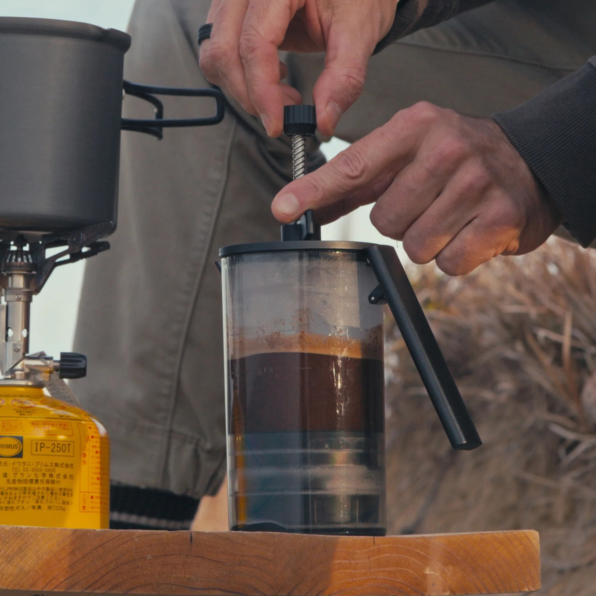 Person pressing down the plunger of a black-handled French press with dark coffee inside on a wooden surface.
