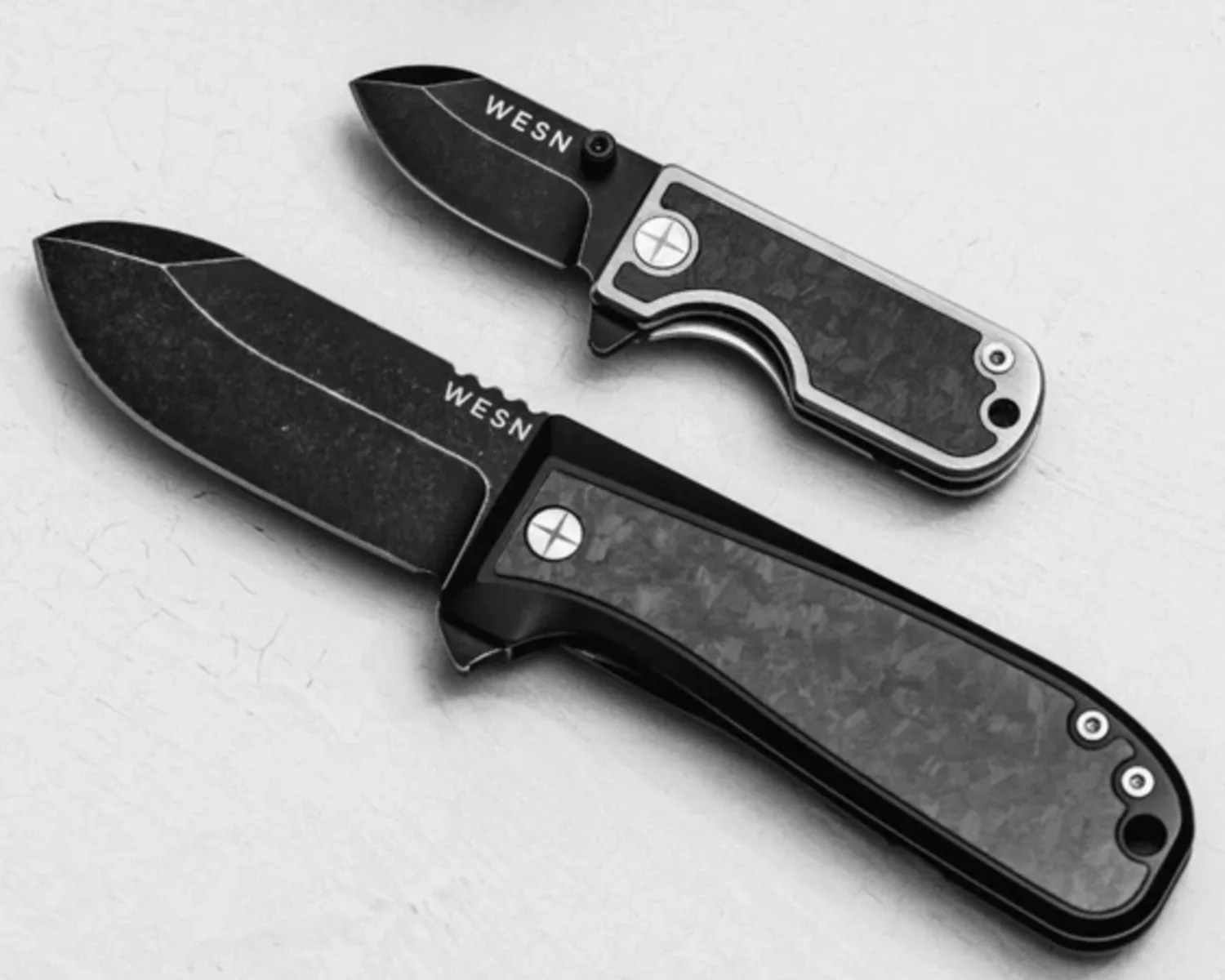 Two black folding knives with textured handles and blades marked "WESN" on a light surface.