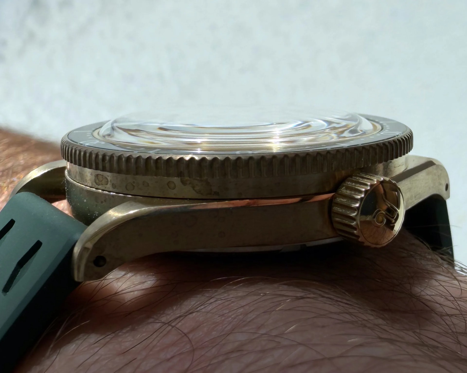 Close-up of a bronze watch case with a domed crystal and a green rubber strap on a wrist.