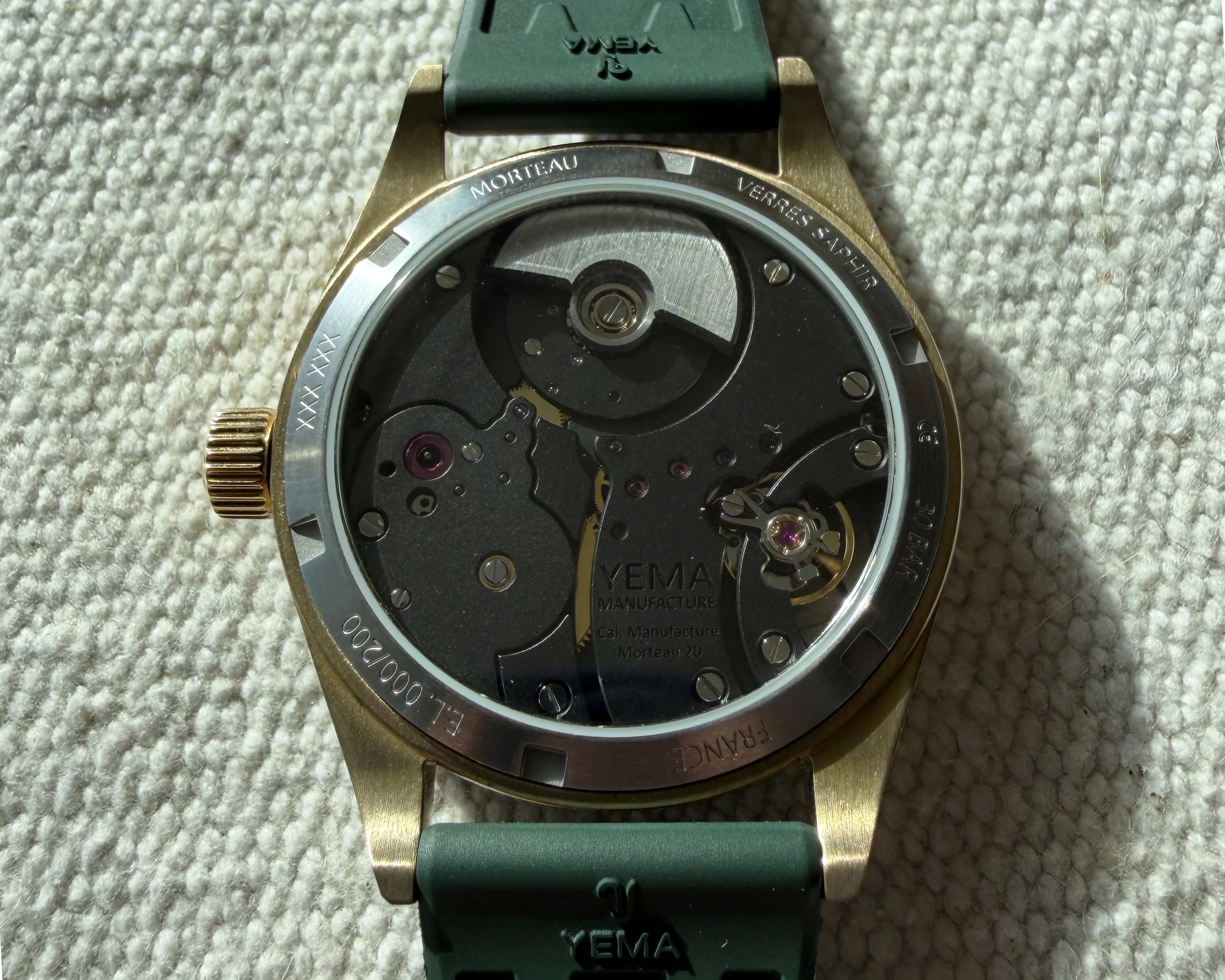 Back of a Yema wristwatch with visible mechanical movement, gold case, and green strap on a textured fabric surface.