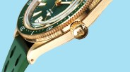Close-up of a gold watch case with a green bezel and dial, featuring a green rubber strap and a gold crown with an engraved logo.