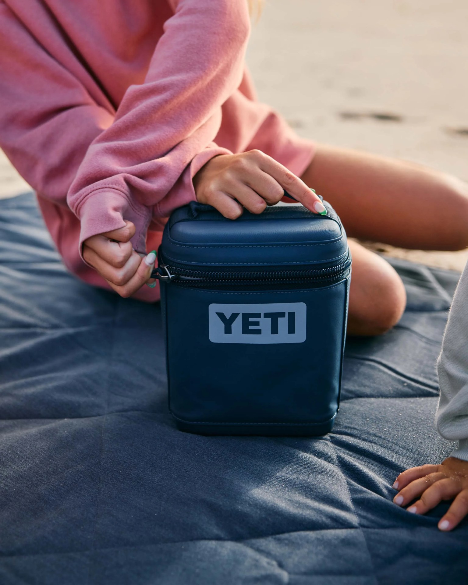 Navy blue YETI soft cooler being held by a person in a pink sweatshirt sitting on a dark quilted blanket.