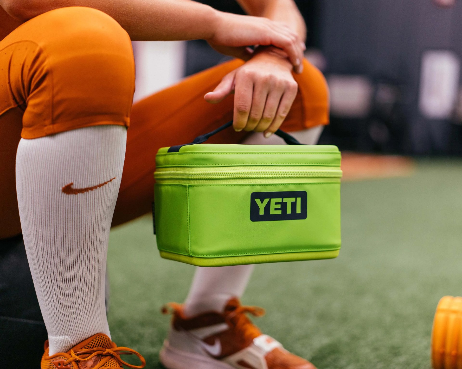 Person holding a lime green YETI cooler bag with black handle, wearing orange pants, white Nike socks, and orange sneakers.