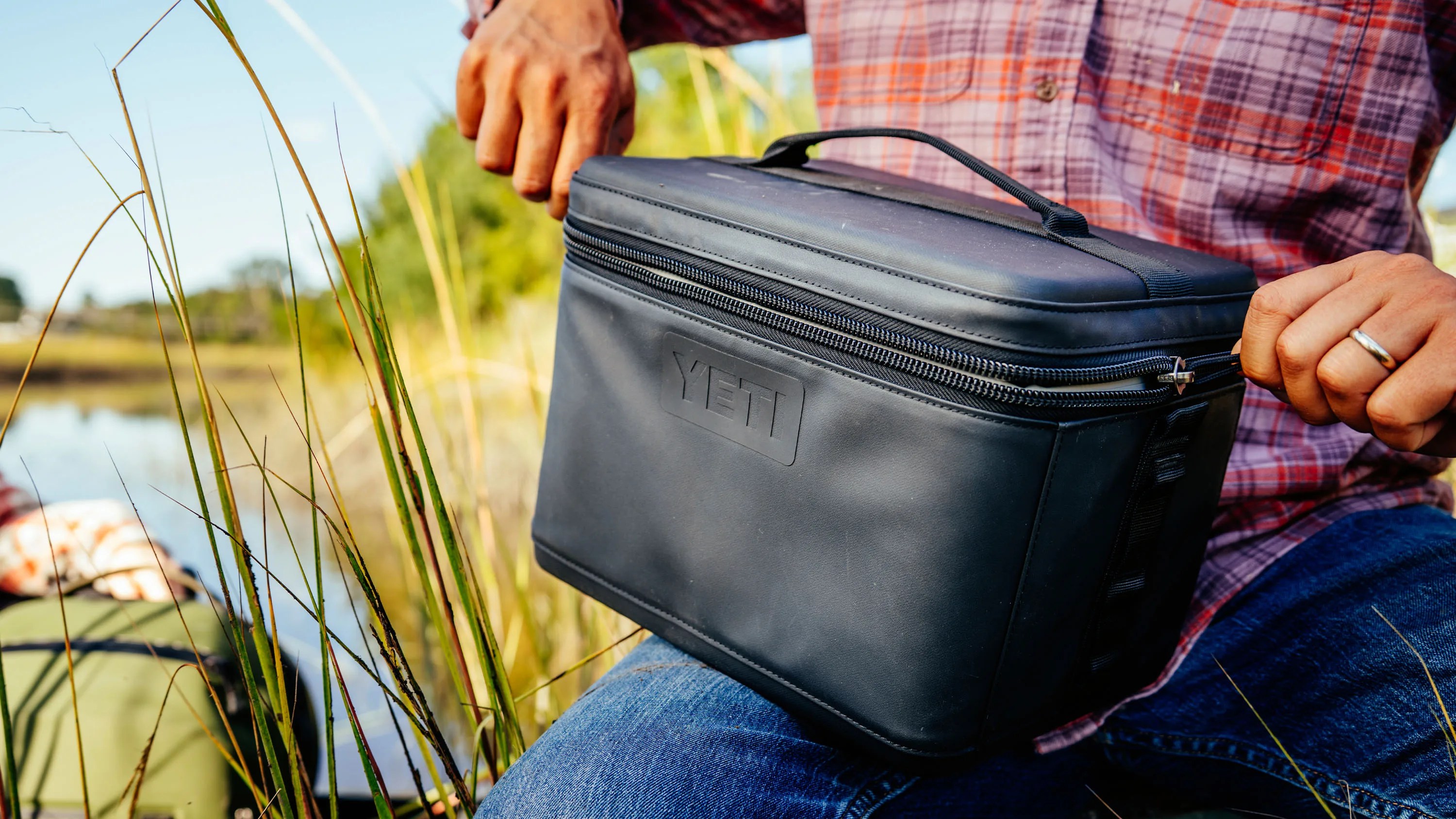 Black YETI soft cooler bag with zipper held by a person wearing a red plaid shirt and jeans near tall grass.
