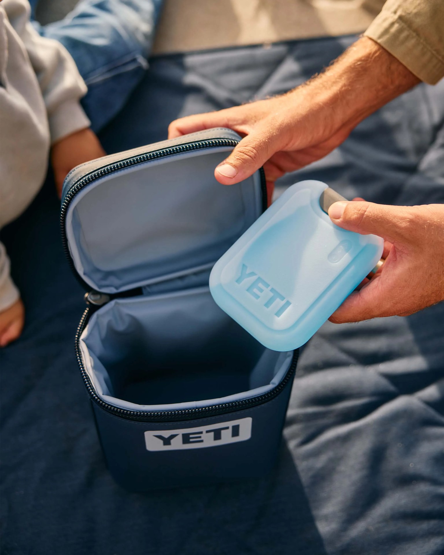 Blue YETI cooler bag with a matching blue ice pack held above it on a dark fabric surface.