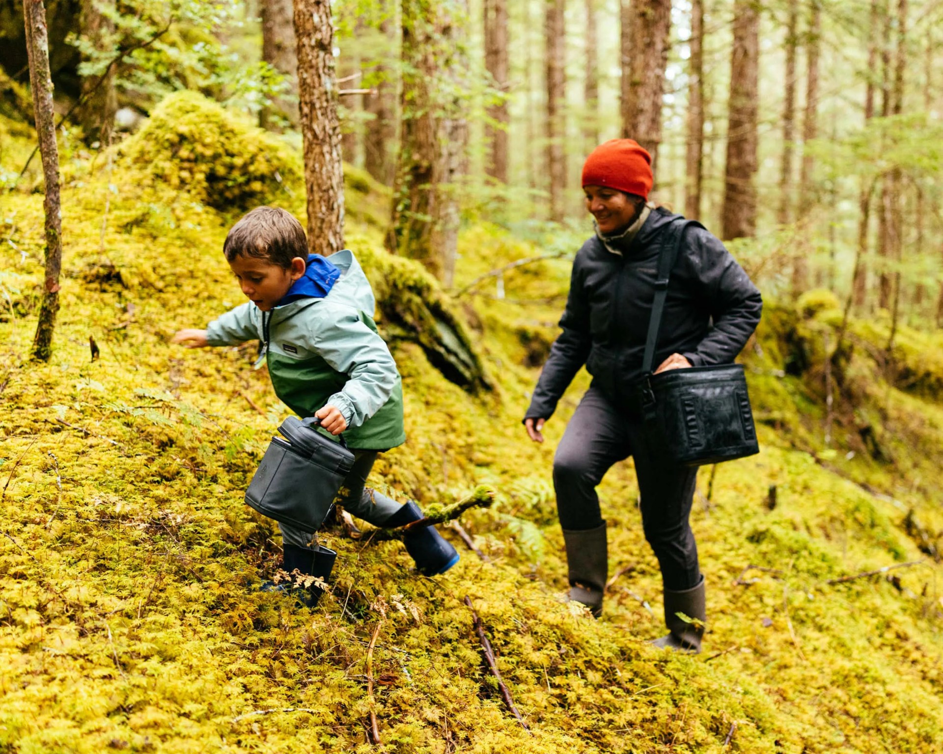 Child in a green jacket and blue boots climbing a mossy hill with a black bucket, followed by a person in a red beanie and dark jacket carrying a black bag.