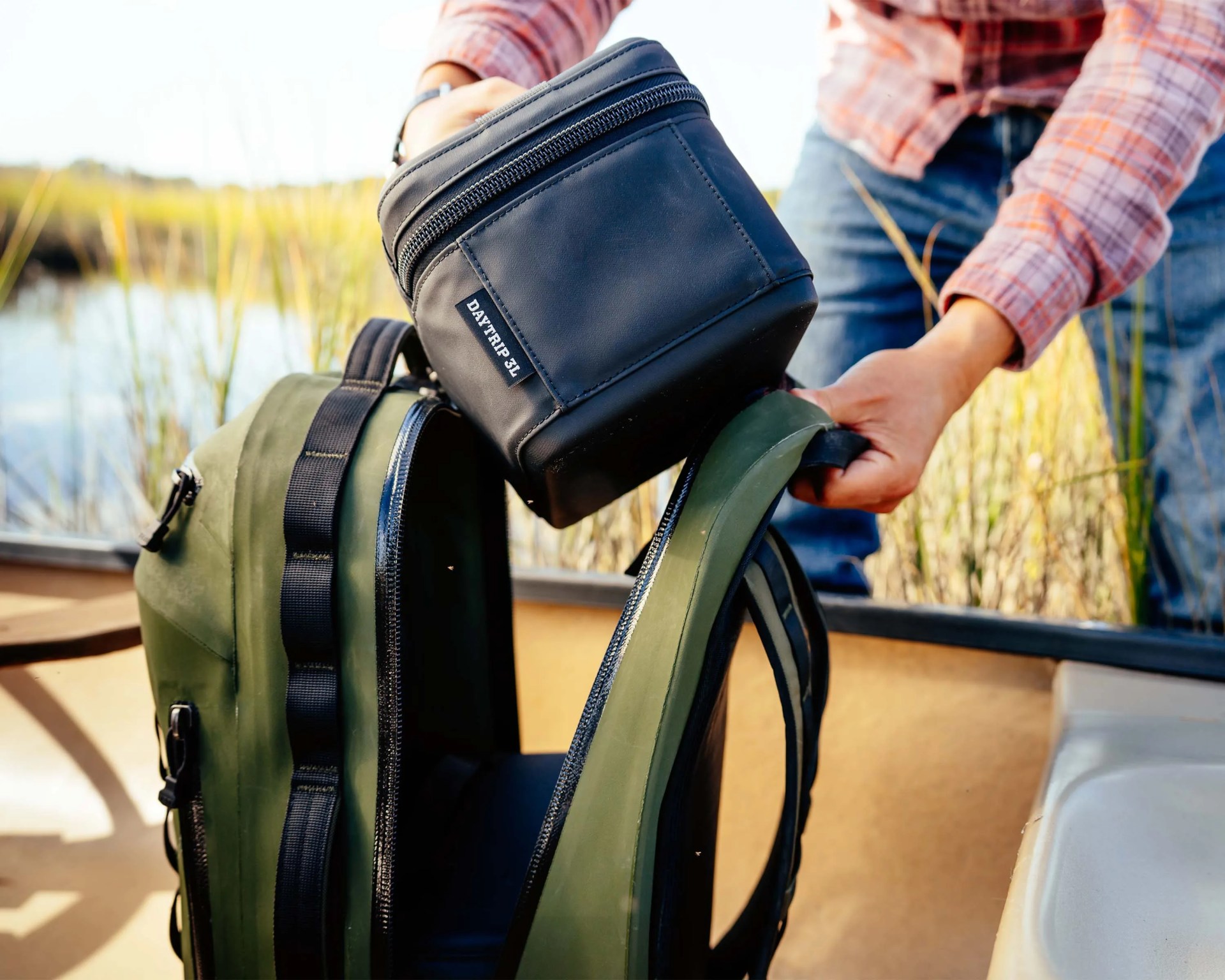 Person placing a black Daytrip 3L bag into an open olive green backpack in a boat.