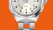 Close-up of a silver DEFY Swiss-made wristwatch with a date window and orange accent on the second hand against an orange background.