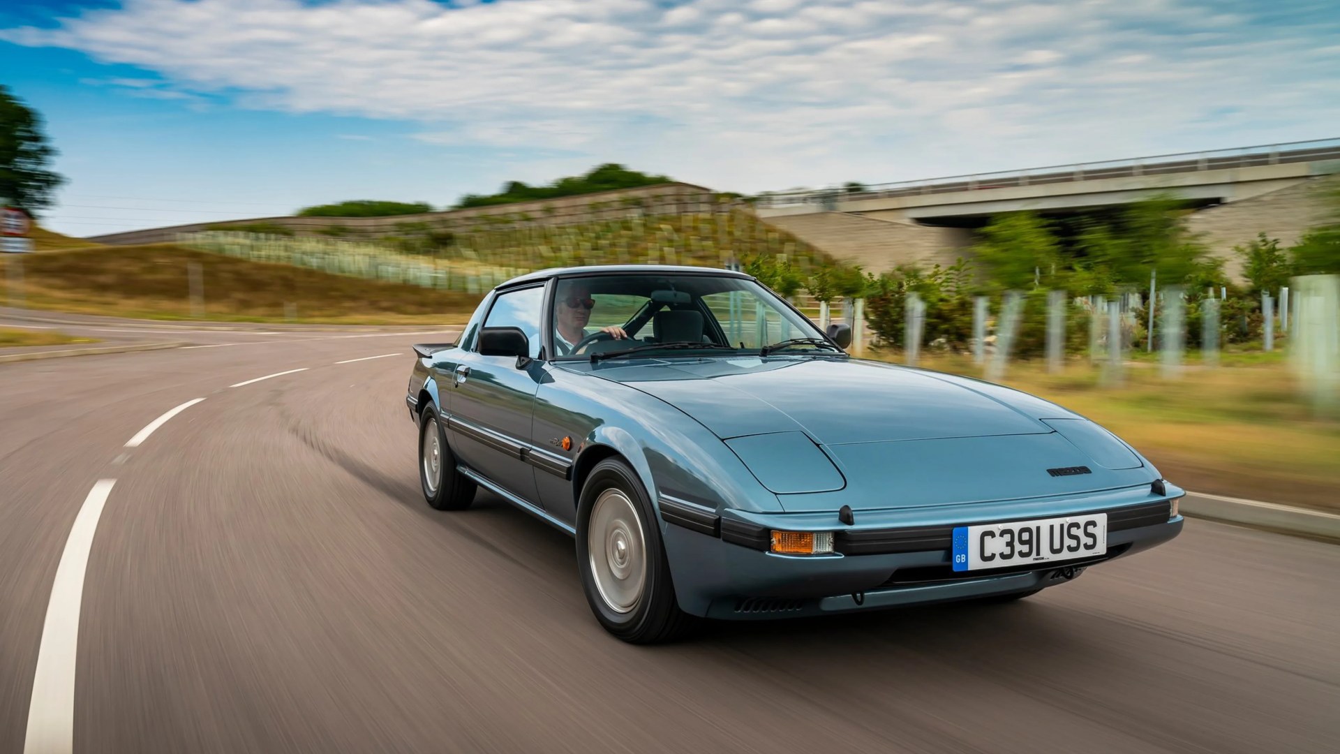 Blue classic Mazda RX-7 coupe driving on a curved road with motion blur.