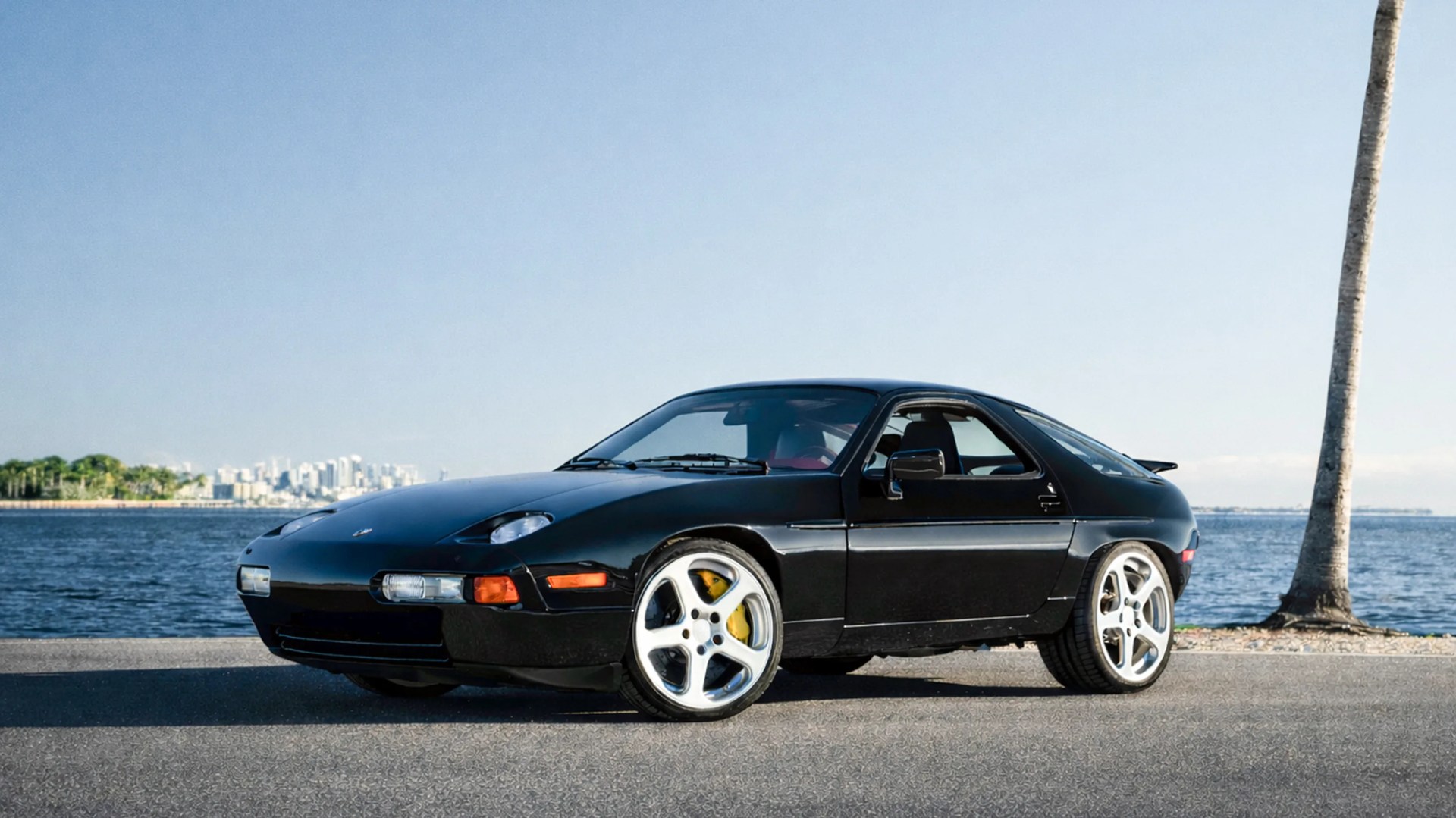 Black vintage sports car with silver five-spoke wheels parked on a waterfront road near a palm tree.