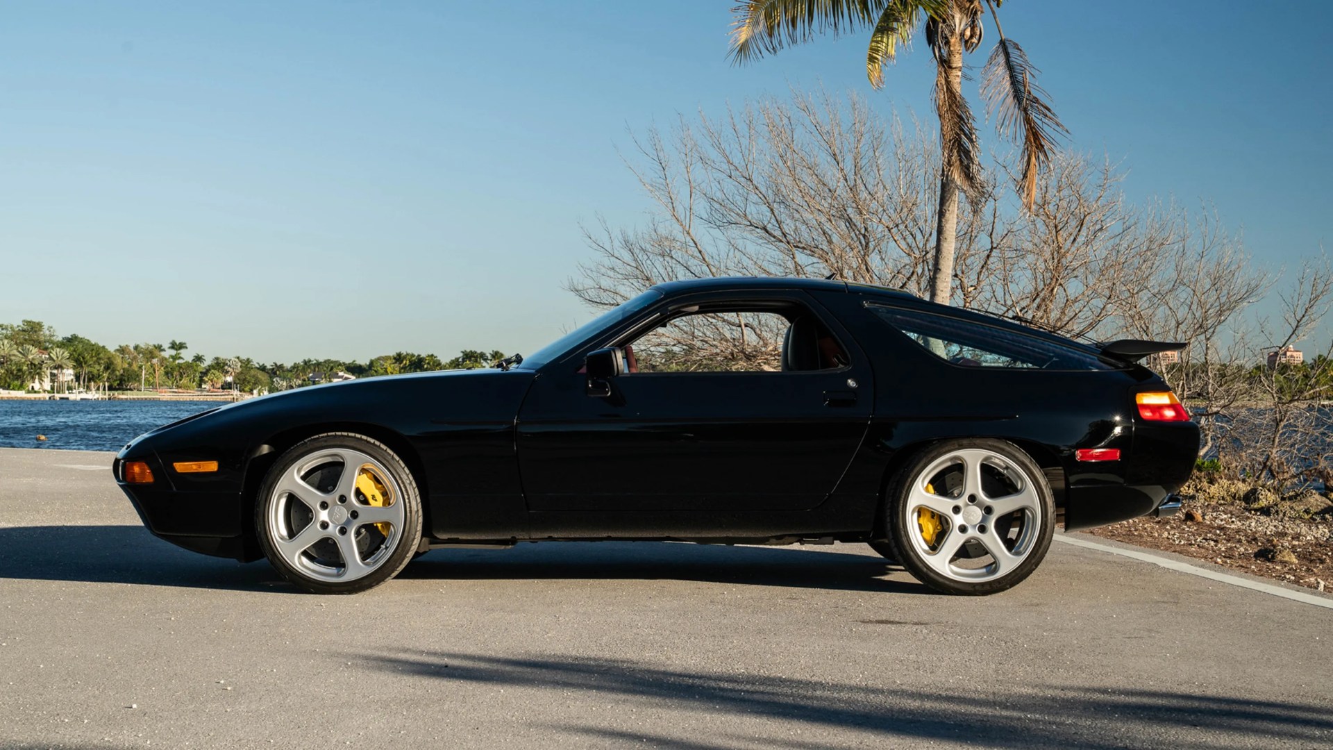 Black classic sports car with silver five-spoke wheels and yellow brake calipers parked near water.