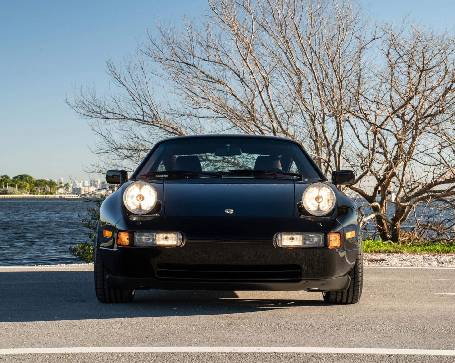 Black Porsche 928 with pop-up headlights on by a waterfront road with leafless trees in the background.