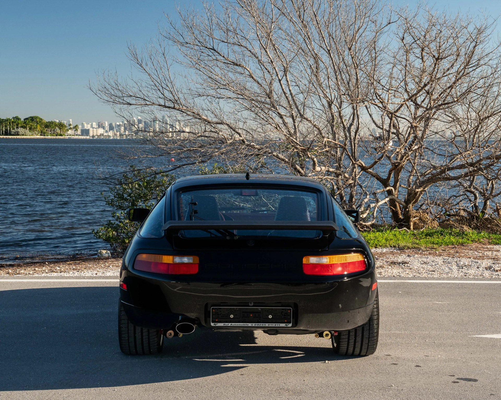 Rear view of a black Porsche sports car parked on a road near a body of water with leafless trees in the background.