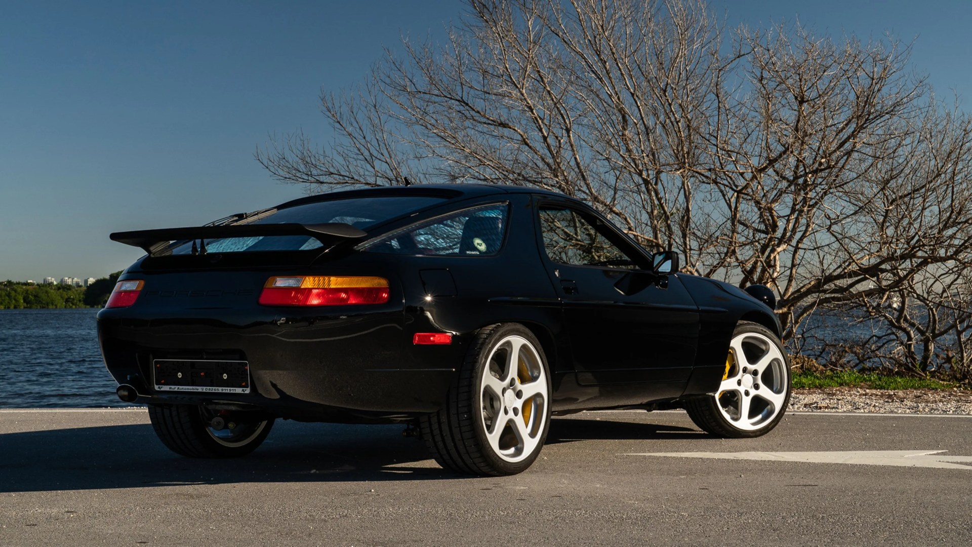 Black Porsche 928 sports car with white wheels parked near a body of water and leafless trees.