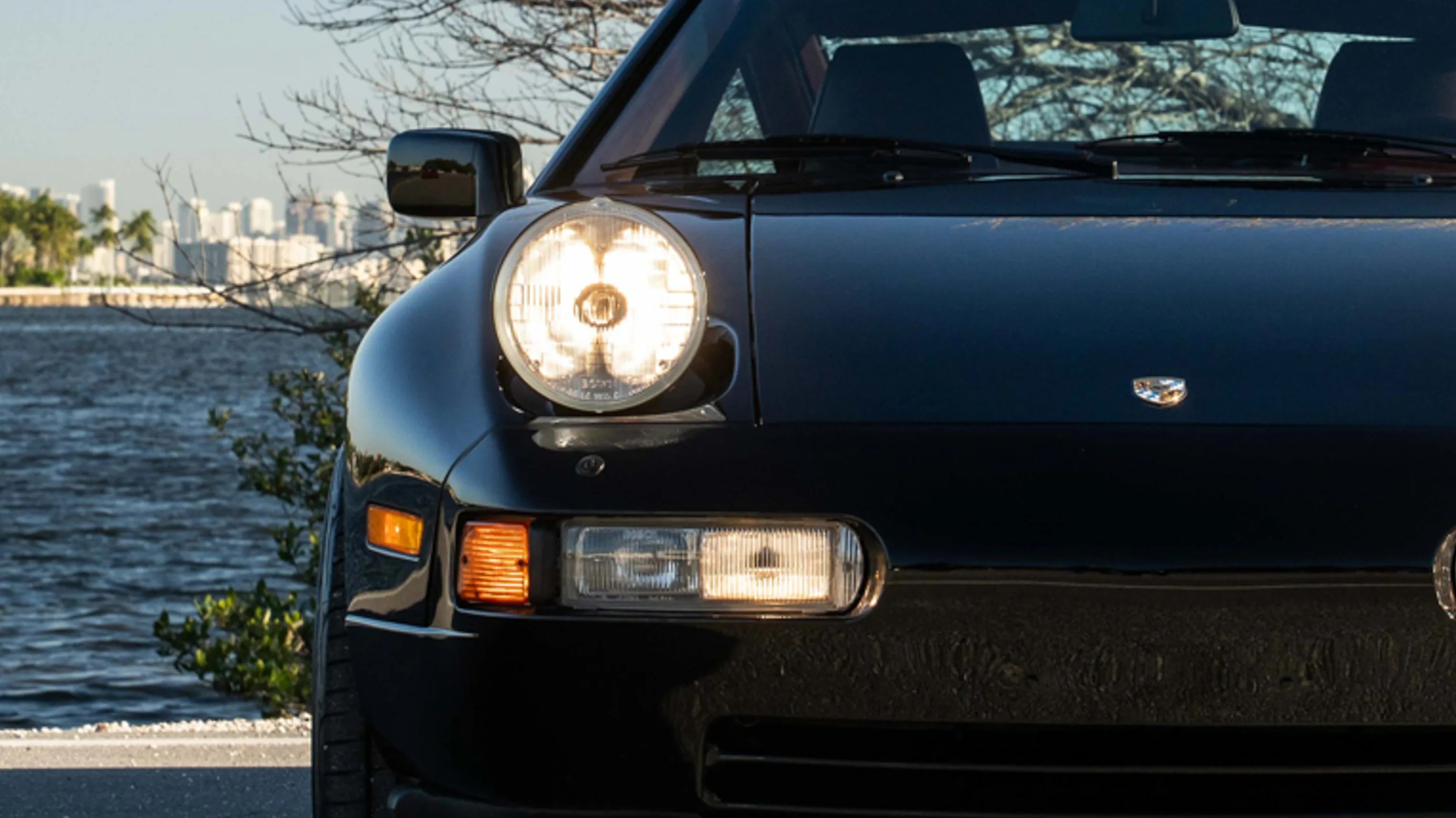 Front left side of a black Porsche 911 with headlights on near a waterfront.