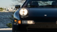 Front left side of a black Porsche 911 with headlights on near a waterfront.