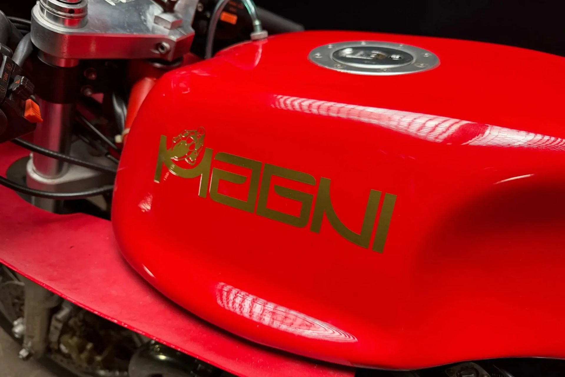 Glossy red motorcycle fuel tank with gold "MAGNI" logo and silver fuel cap.