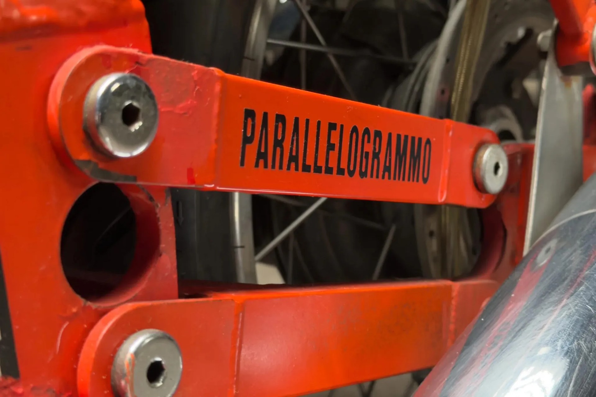 Close-up of an orange metal part with bolts and the word "PARALLELOGRAMMO" printed in black.