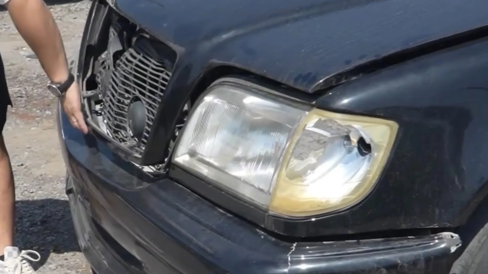 Front of a black car with a broken right headlight and damaged grille, with a person&rsquo;s hand near the grille.