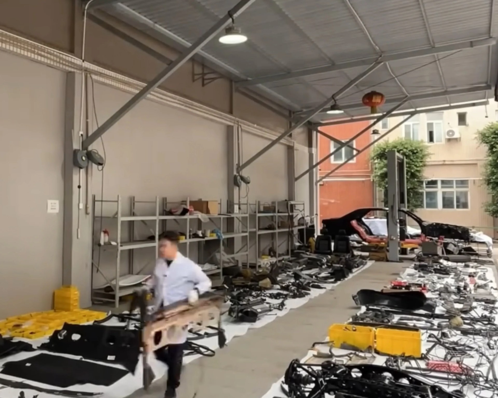 Workshop with car parts laid out on the floor and shelves, a person in a white coat carrying a part.