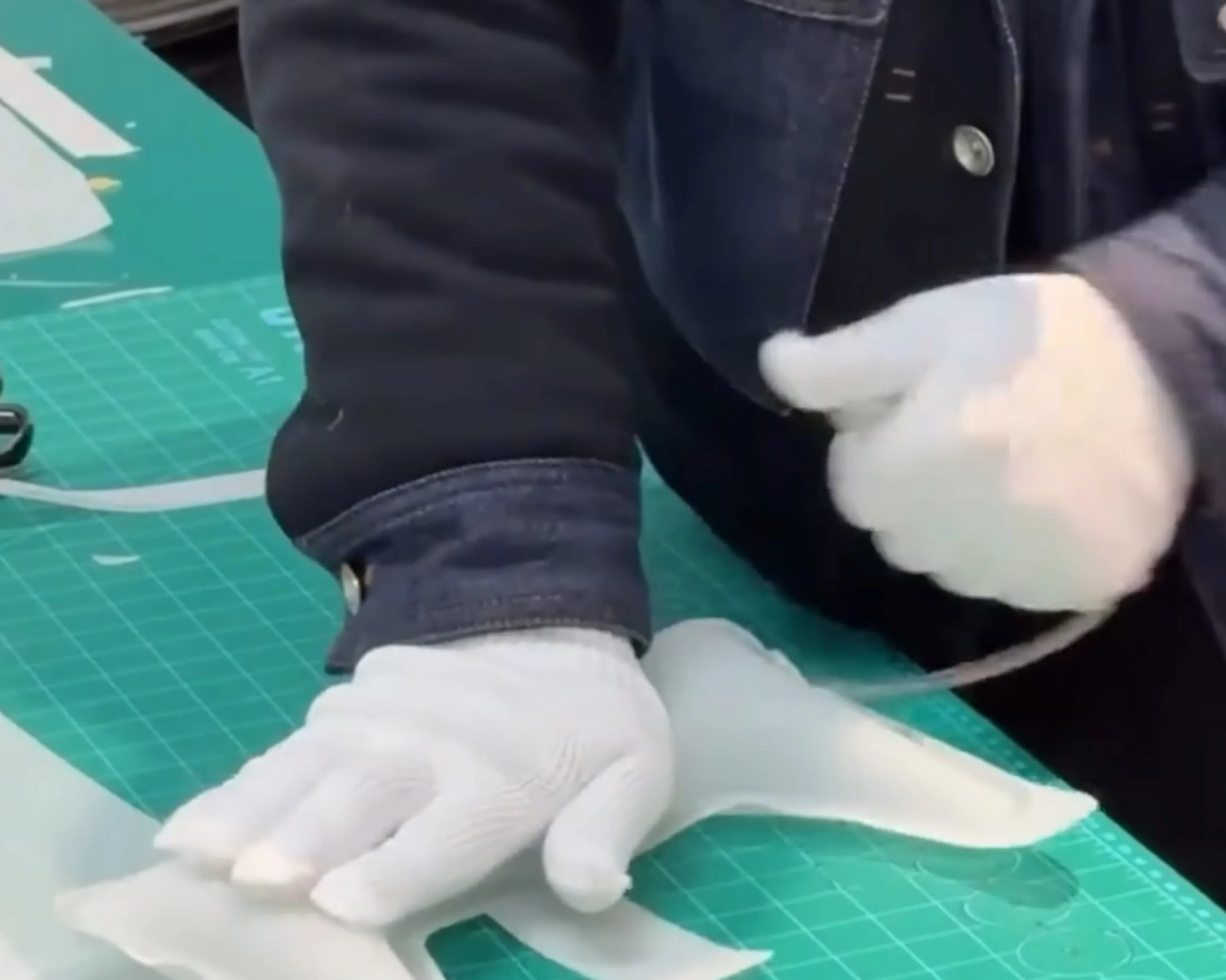 Person wearing white gloves and a dark jacket working on a translucent plastic sheet on a green cutting mat.