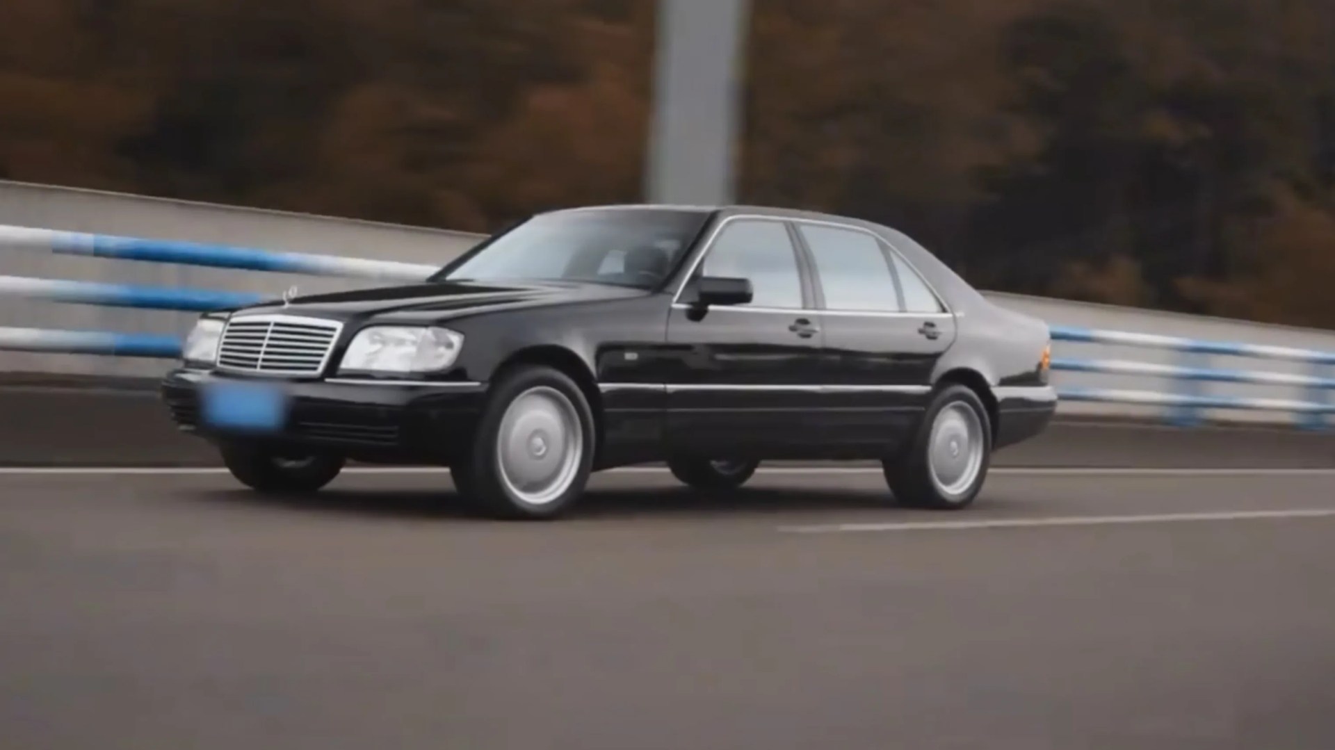 Black Mercedes-Benz sedan driving on a road with blue and white guardrails in the background.